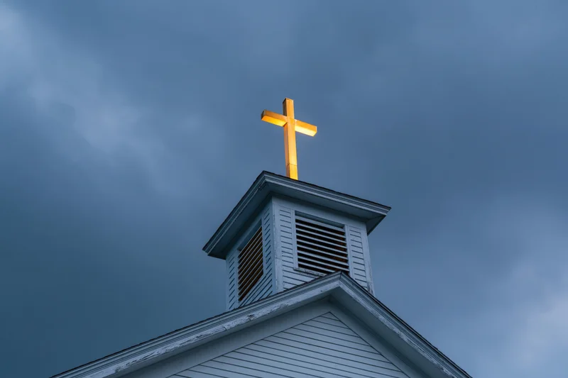 Illuminated cross atop white wooden church steeple against cloudy evening sky