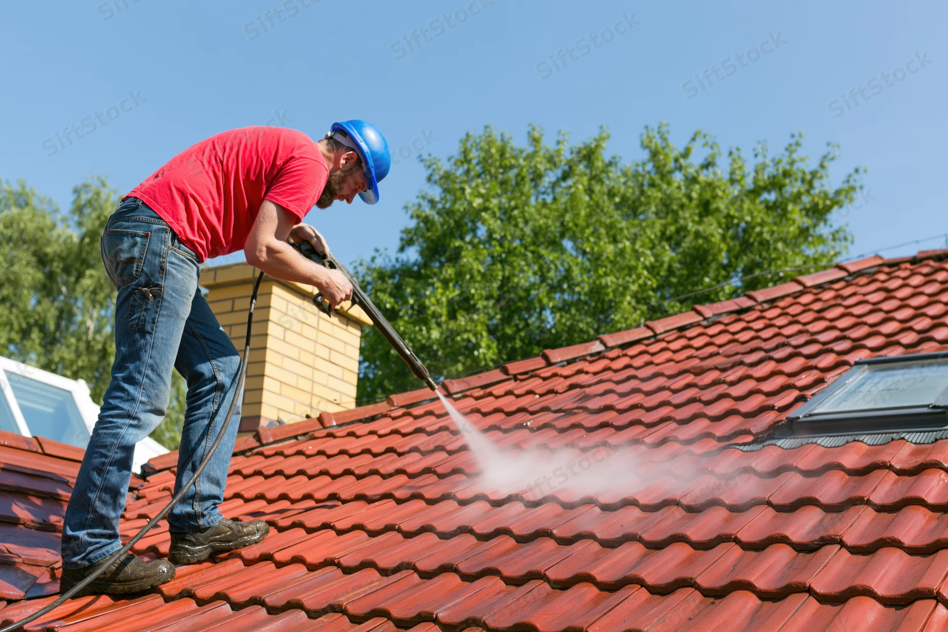 Man pressure washing a red tiled roof outdoors wearing a blue hard hat and red shirt