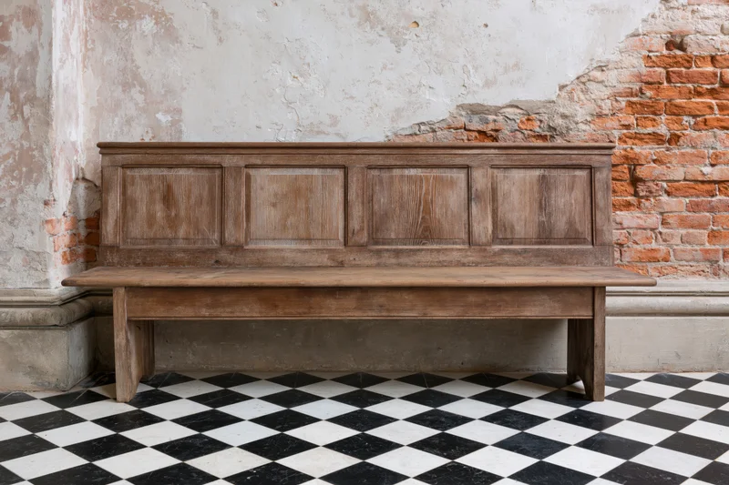 Wooden church pew with paneled backrest against weathered brick and plaster wall on checkered tile floor