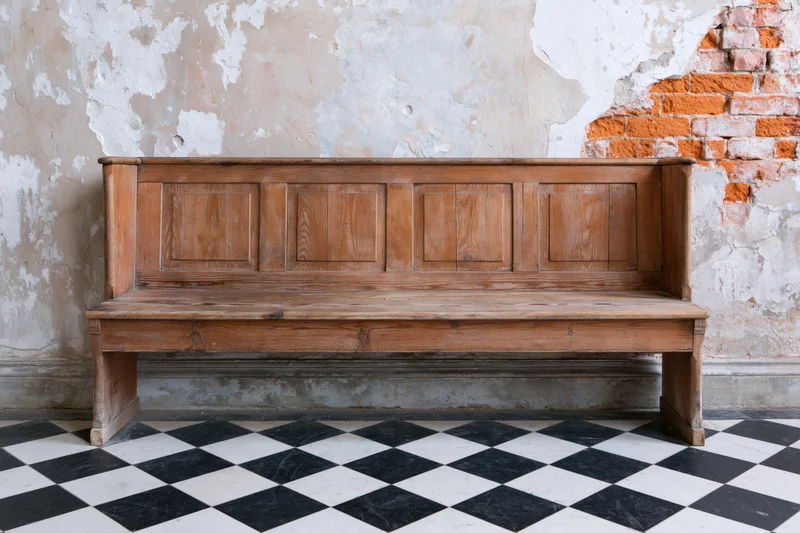 Wooden church pew with paneled backrest against peeling plaster and exposed brick wall on checkered tile floor