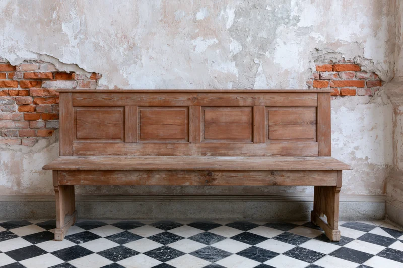 Worn wooden church pew with paneled backrest against peeling plaster and exposed brick wall on checkered tile floor
