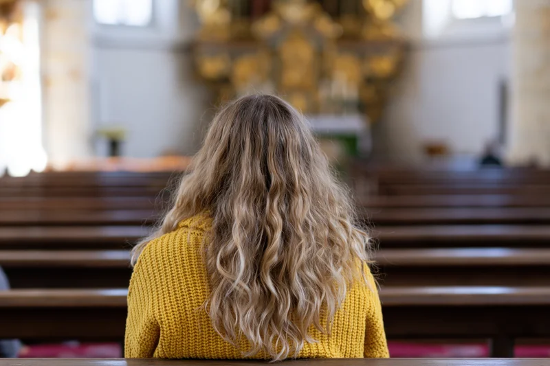 Woman with long wavy blonde hair sitting alone on a church bench facing the altar