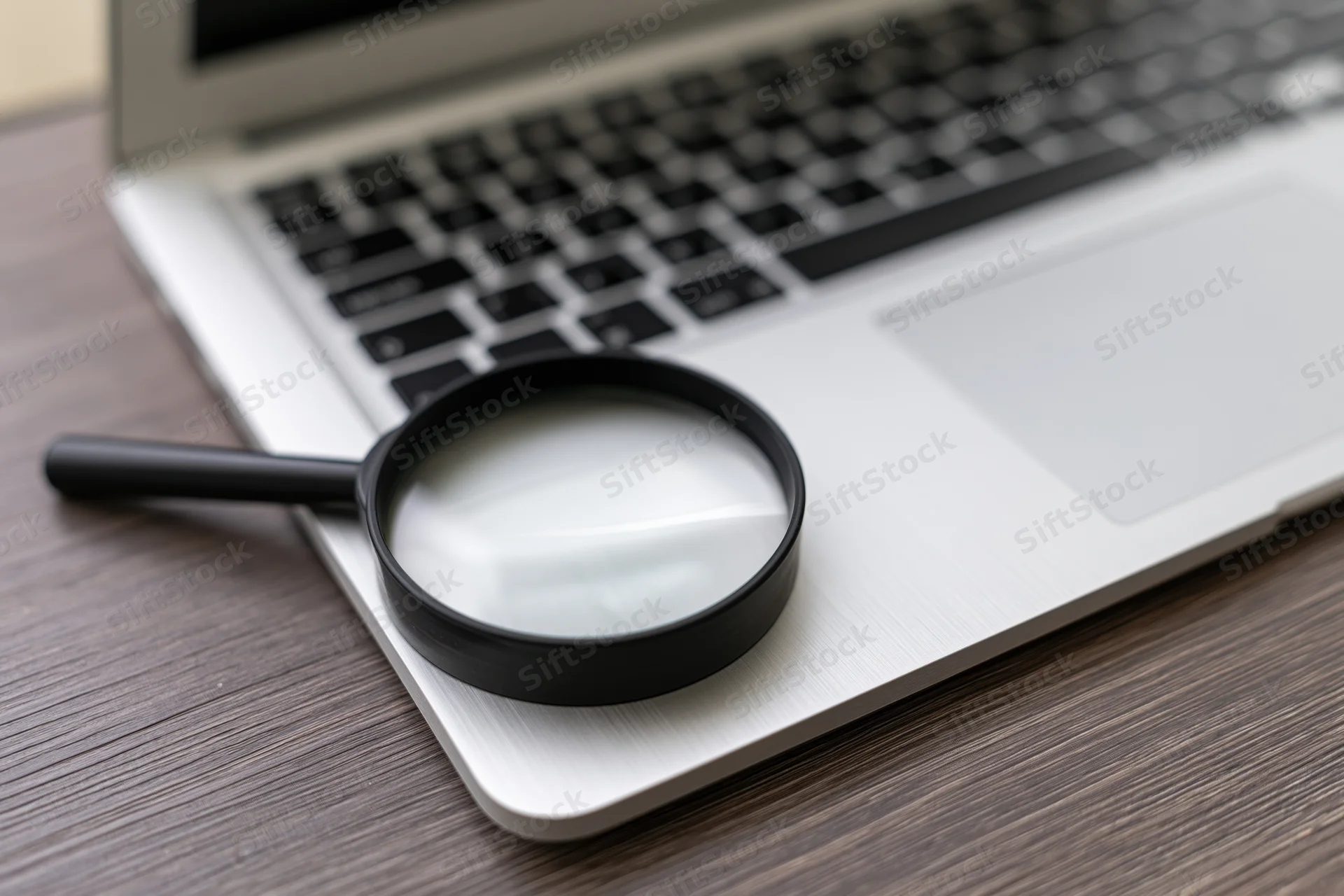 Black magnifying glass resting on open silver laptop keyboard on wooden desk