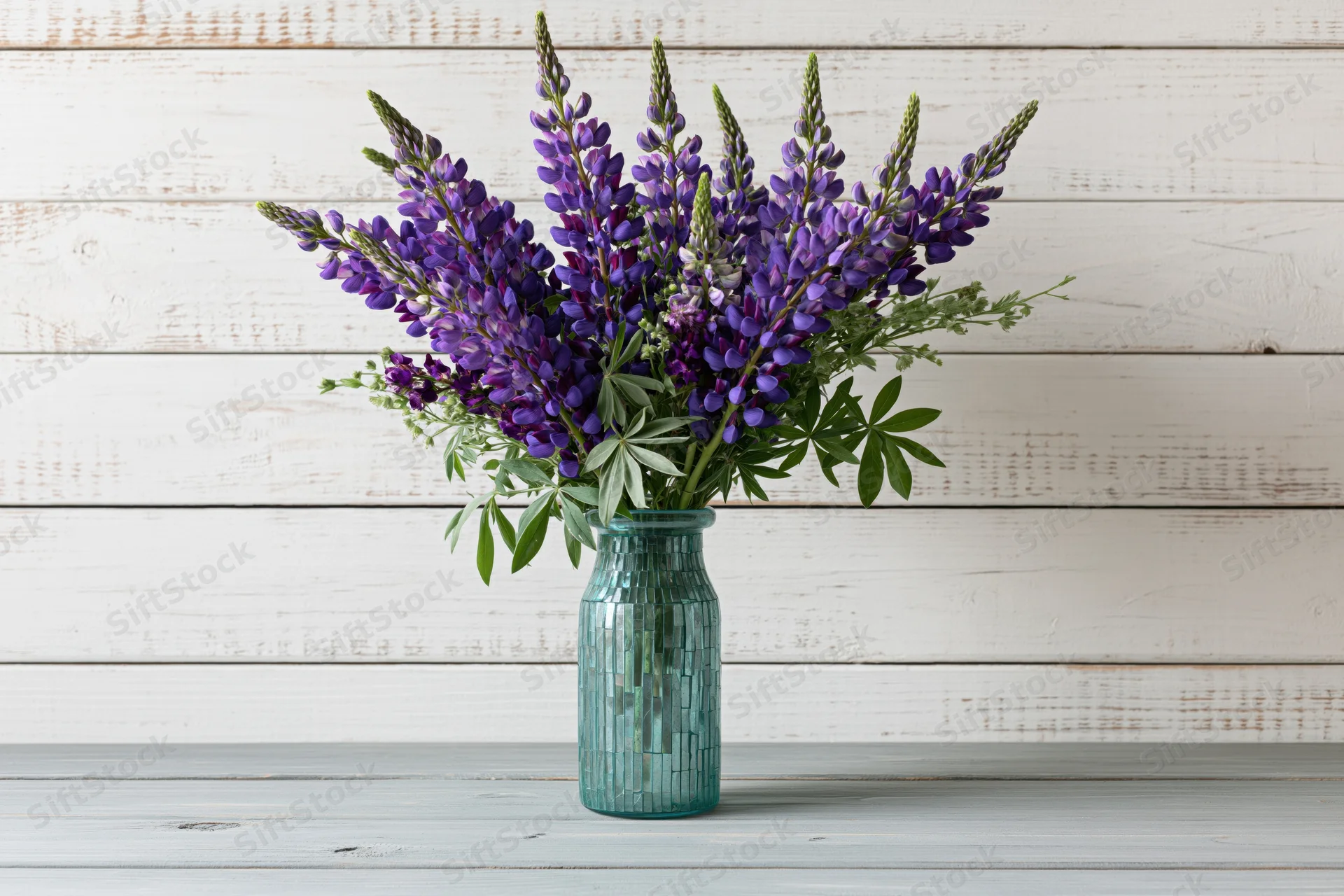 Bouquet of purple and violet lupin flowers with green leaves in a teal mosaic glass vase on a white wooden surface