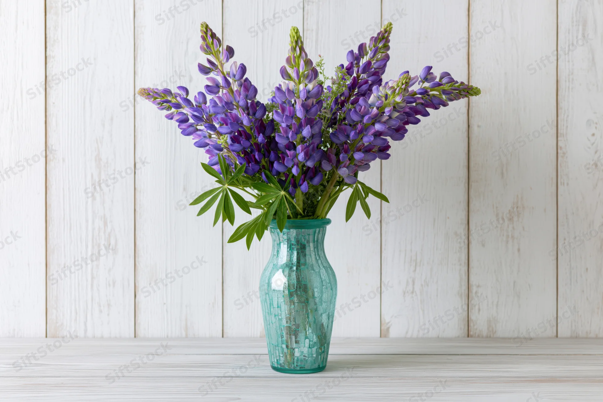 Bouquet of purple and violet lupin flowers with green leaves in a teal mosaic glass vase on a white wooden surface