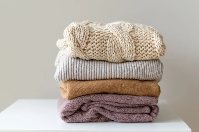 Stack of four folded knit sweaters in neutral tones on white surface