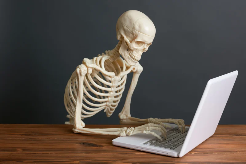 Plastic human skeleton model typing on laptop at wooden desk