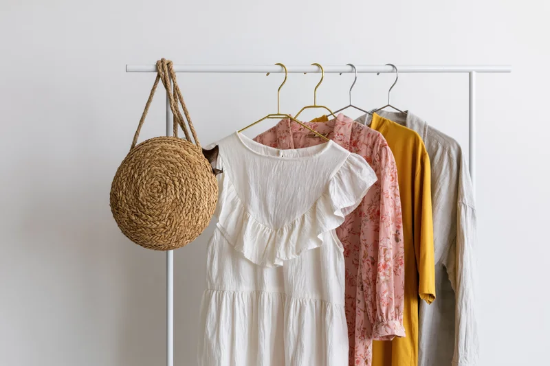 Women's dresses and a straw bag hanging on a white portable clothing rack against a plain wall