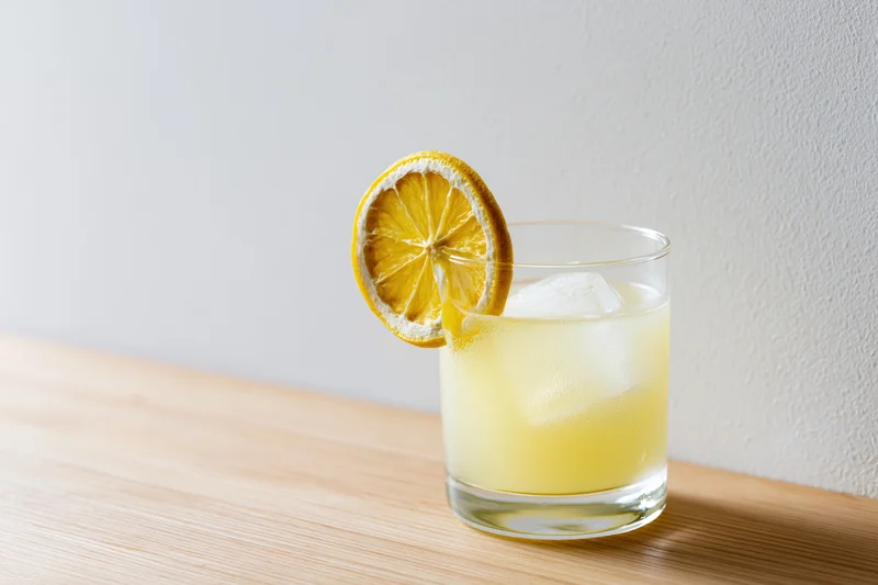 Pale yellow lemonade in a rocks glass with ice and a lemon slice garnish on a wooden table