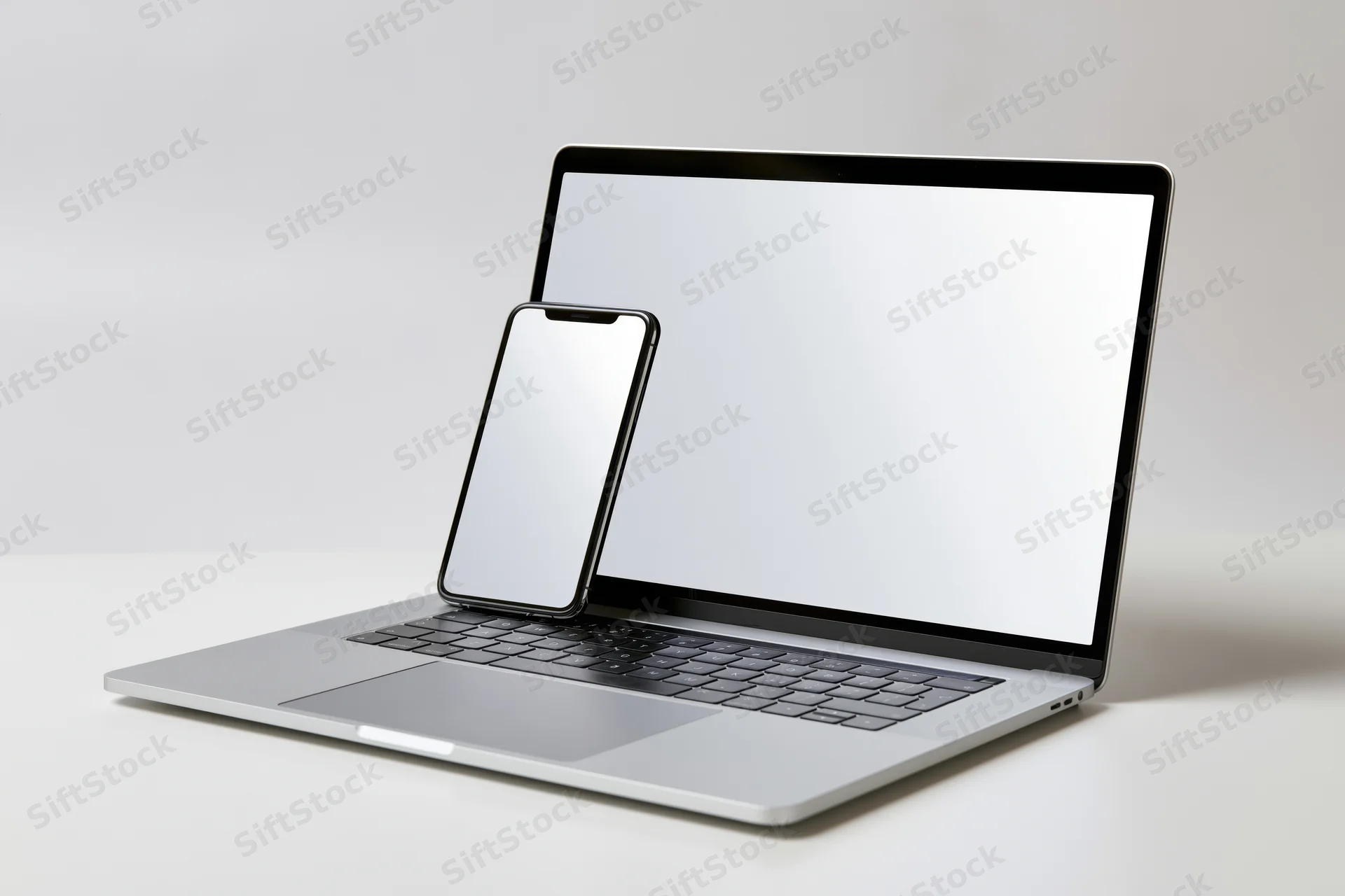 Silver laptop with blank white screen and smartphone on keyboard, studio setup