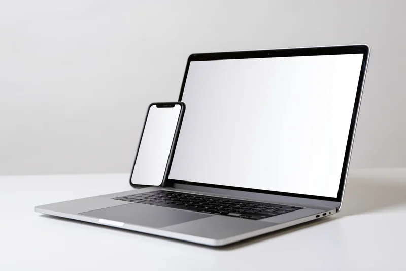 Silver laptop with blank white screen and smartphone beside it on white surface