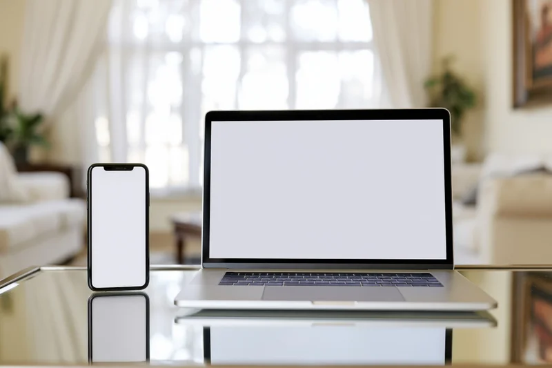 Laptop and smartphone with blank screens on a reflective glass table in a living room