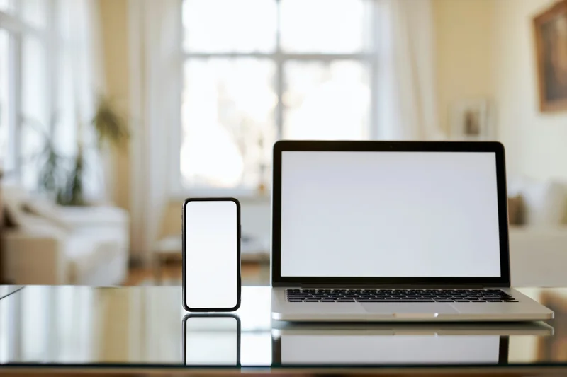 Laptop and smartphone with blank screens on a reflective glass table in a bright home office