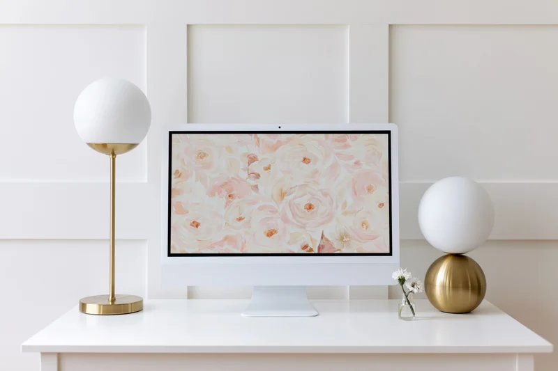 White desktop computer displaying floral wallpaper on a minimalist white desk with gold lamps and small vase of flowers