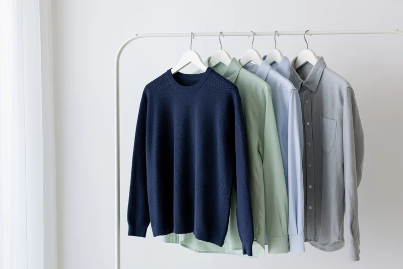 Several garments including a sweater and shirts hanging on a white metal rack in a minimalist interior