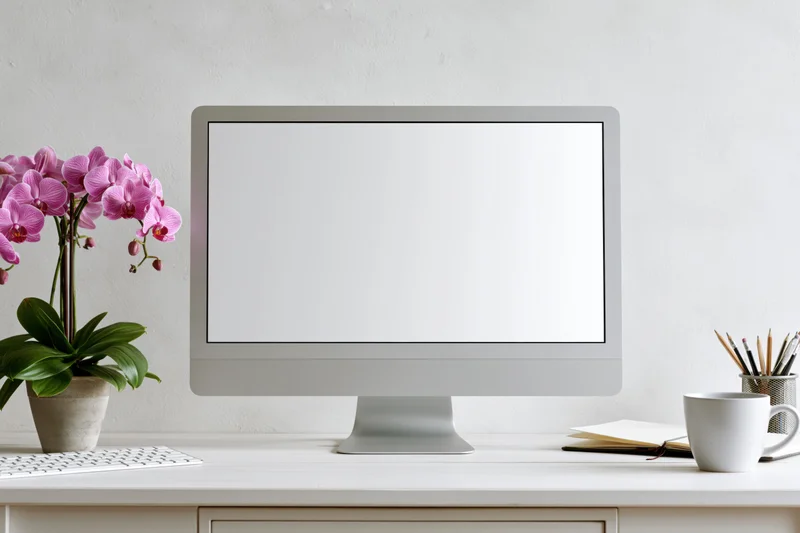 All-in-one computer with blank white screen on a desk beside a pink orchid and office supplies