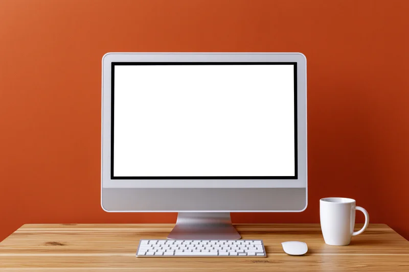 All-in-one desktop computer with blank white screen on wooden desk beside mug and peripherals