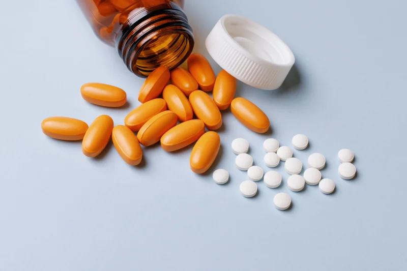 Amber glass medicine bottle lying on its side with orange capsules and white tablets spilled onto a light blue surface