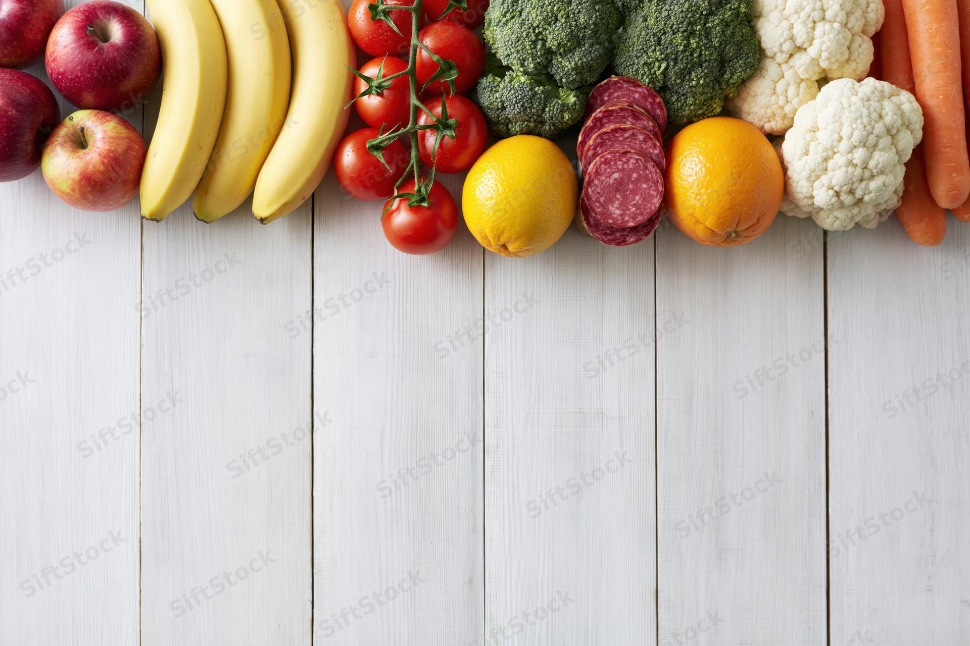 Assorted fresh fruits vegetables and sliced salami arranged along top of white wooden table