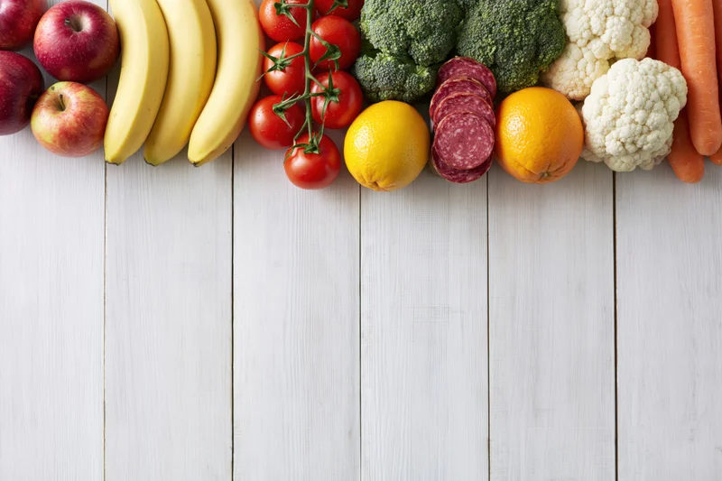 Assorted fresh fruits vegetables and sliced salami arranged along top of white wooden table