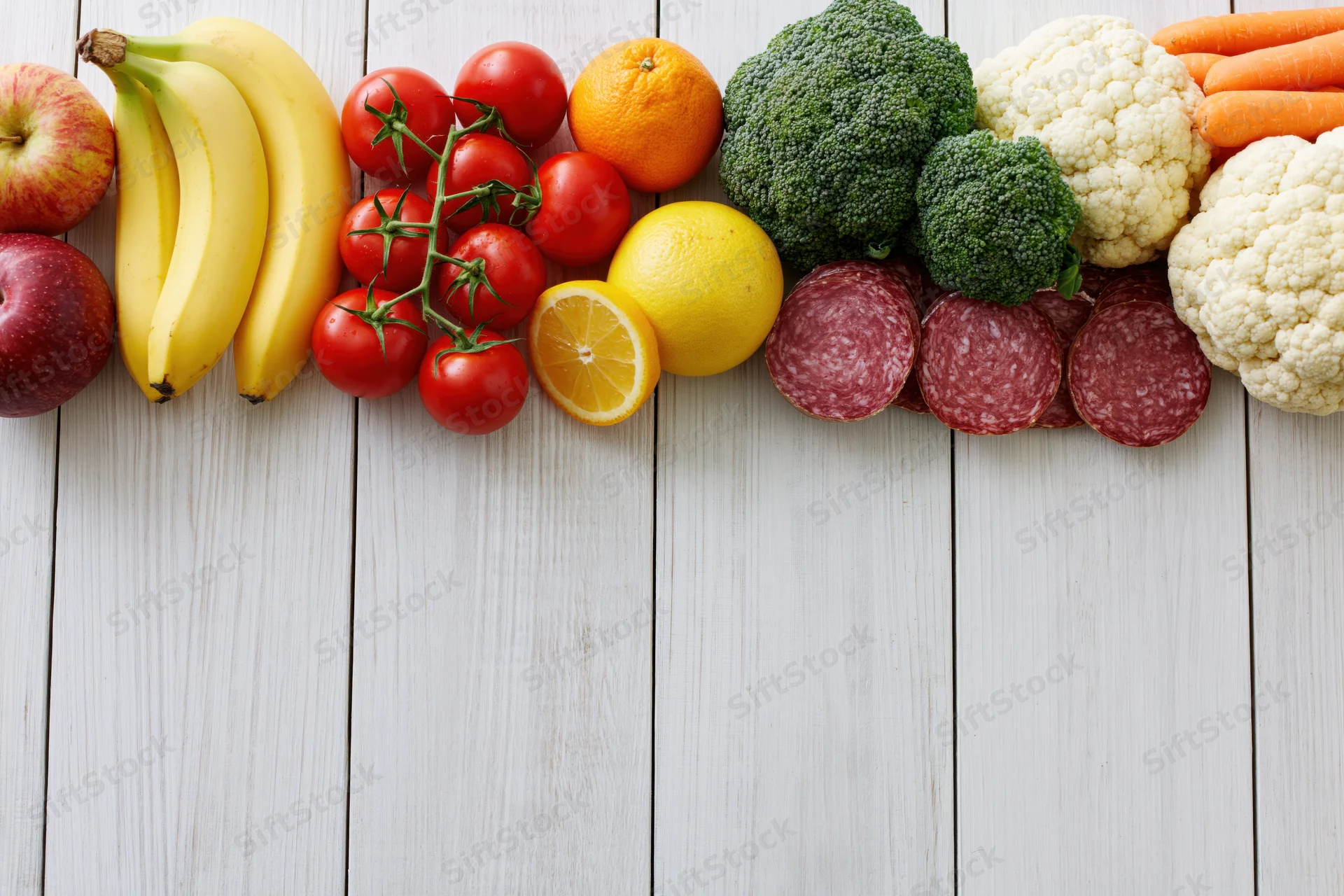 Assorted fresh fruits vegetables and sliced salami arranged in a row on a white wooden surface