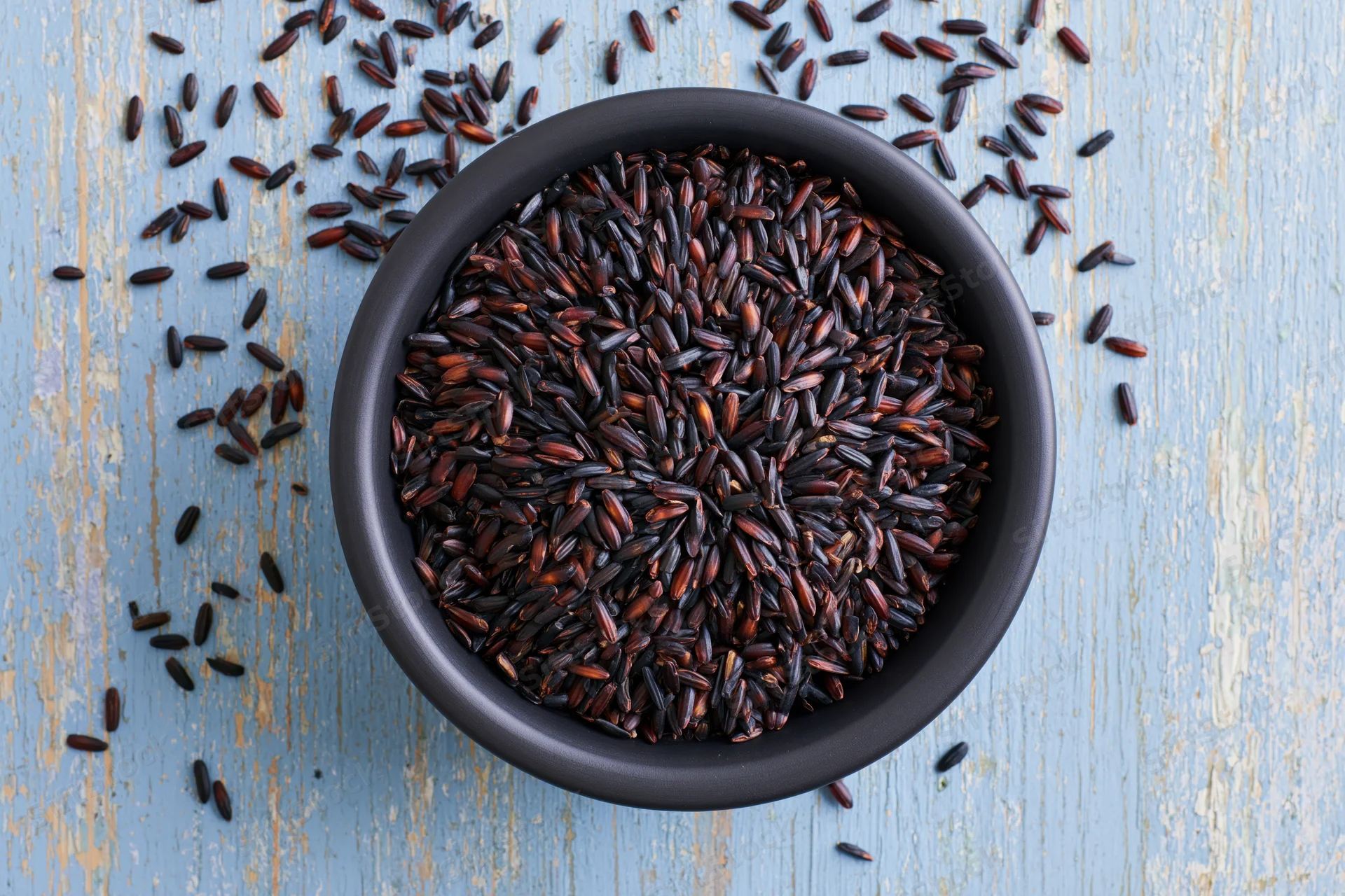 Black rice grains in a matte black ceramic bowl on a blue wooden surface, top view with scattered grains around