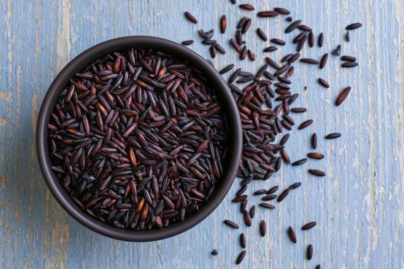 Black rice grains in a matte black ceramic bowl on a blue weathered wooden surface