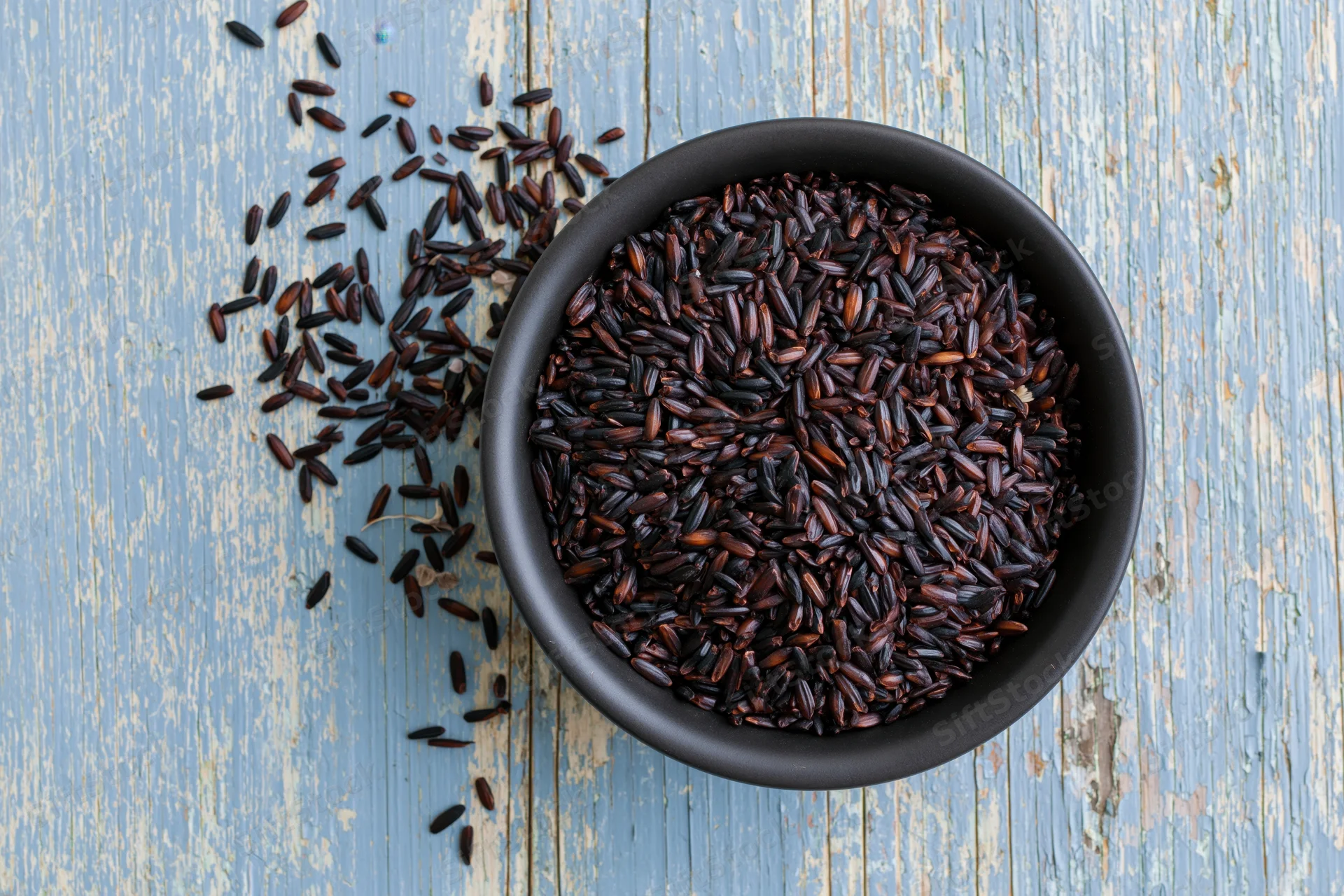 Black rice grains in a matte black ceramic bowl on a blue wooden surface with scattered grains beside it
