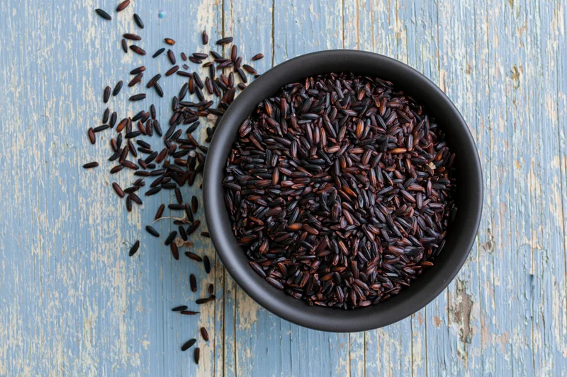 Black rice grains in a matte black ceramic bowl on a blue wooden surface with scattered grains beside it