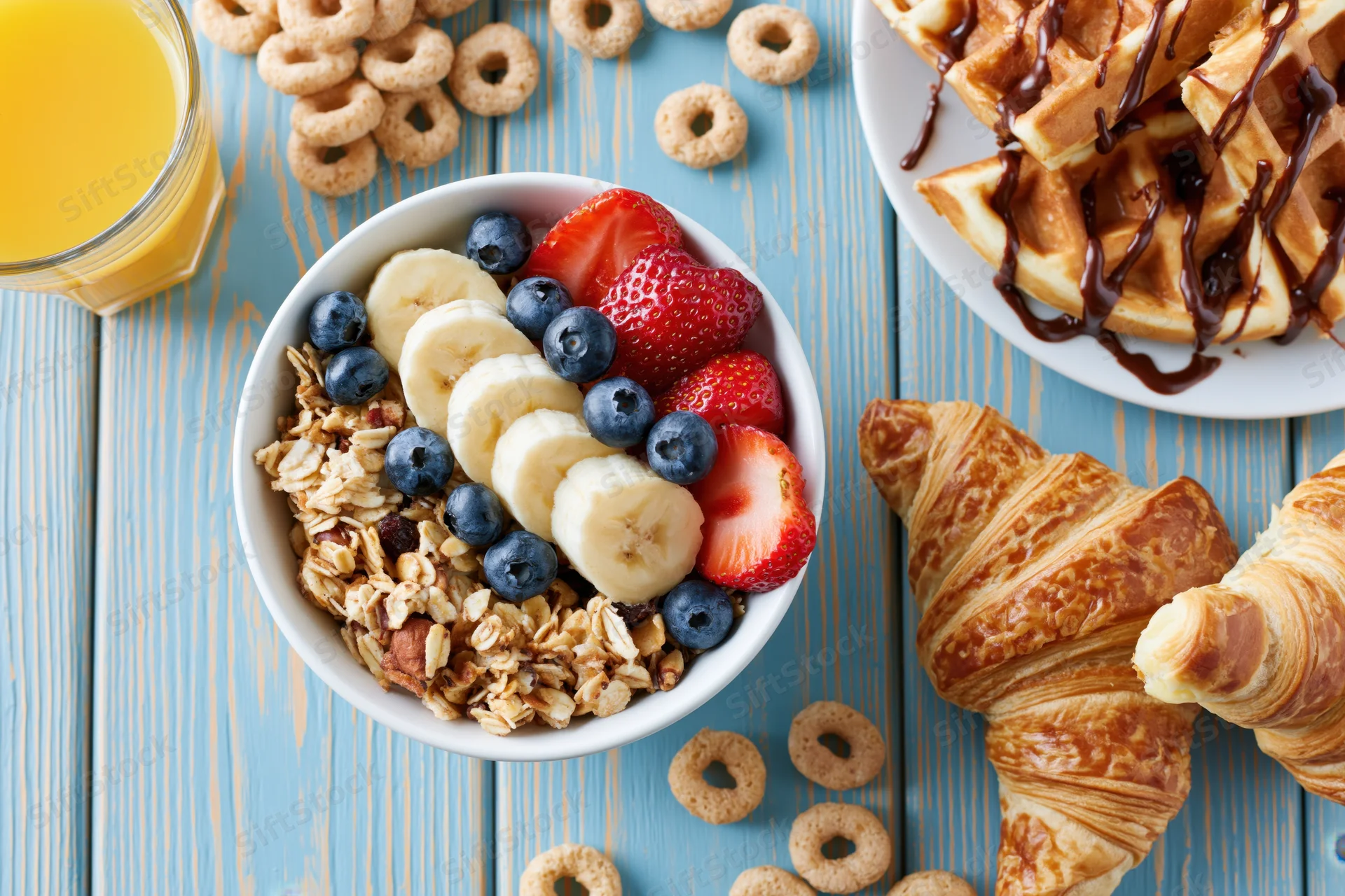 Breakfast flat lay with granola bowl, croissants, waffles, and orange juice on blue wooden table