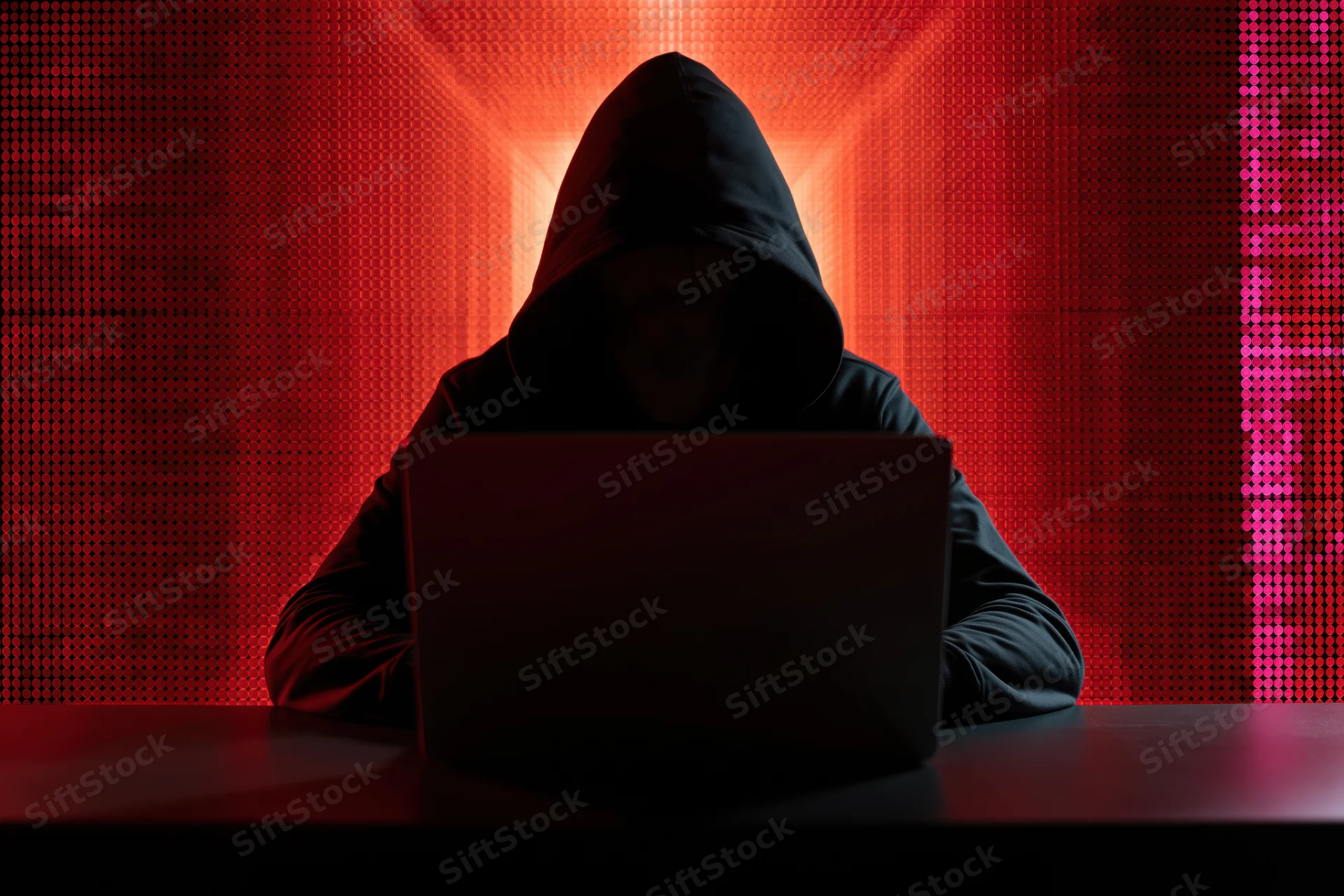 Hooded figure working on laptop in dark room with red digital background
