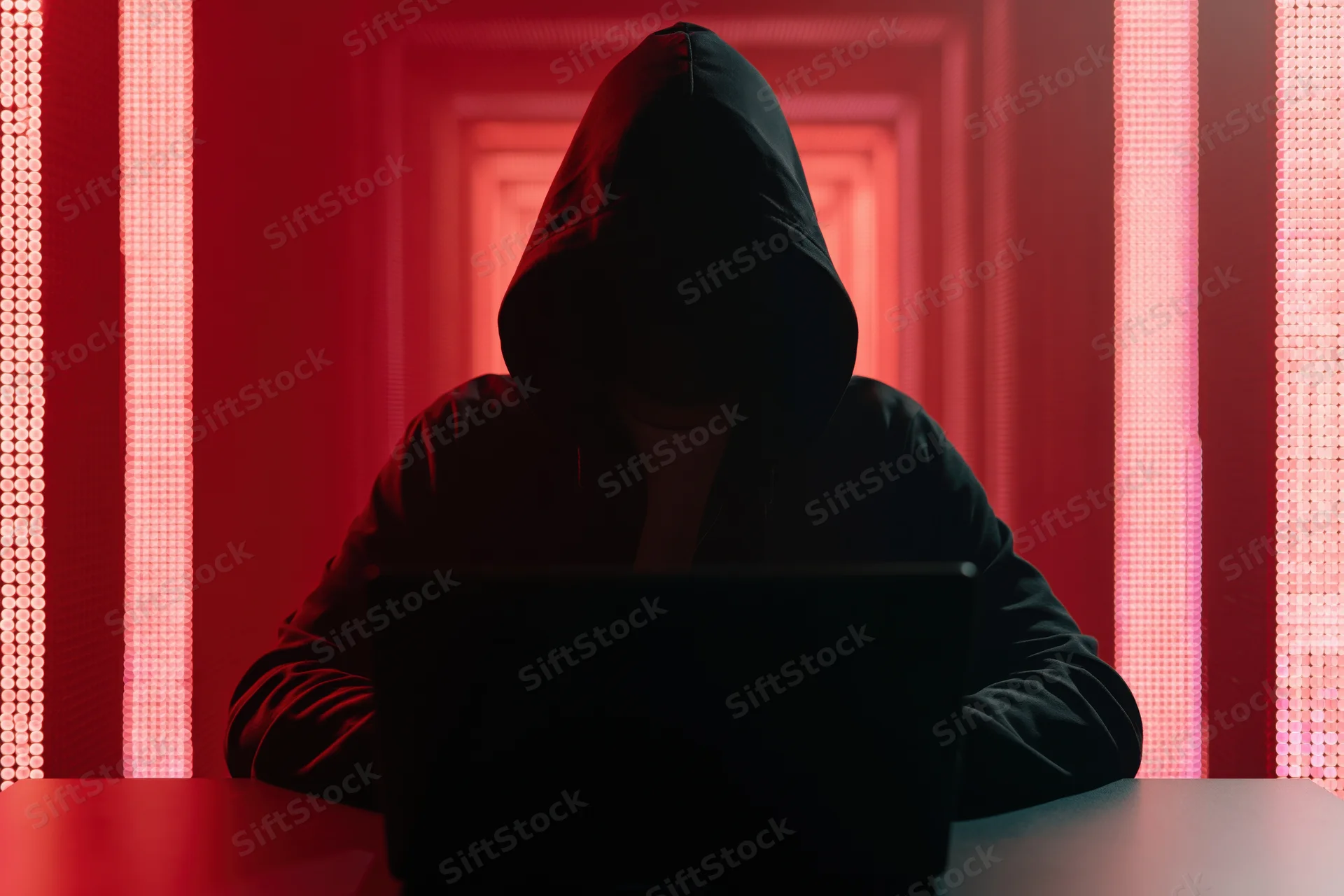 Hooded figure working on laptop in red-lit tunnel setting