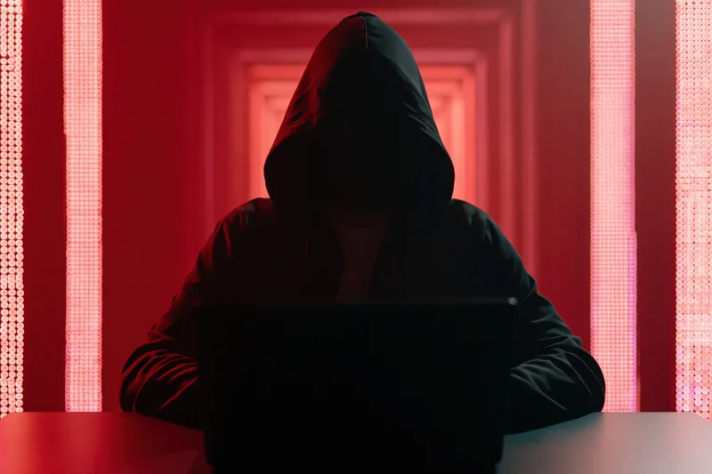 Hooded figure working on laptop in red-lit tunnel setting