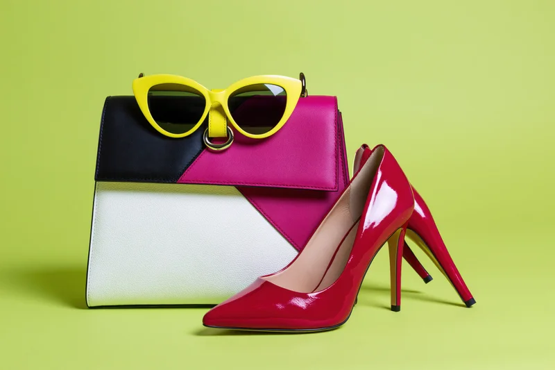 Colorful fashion accessories including a structured handbag, red high heels, and yellow cat-eye sunglasses arranged on a lime green background