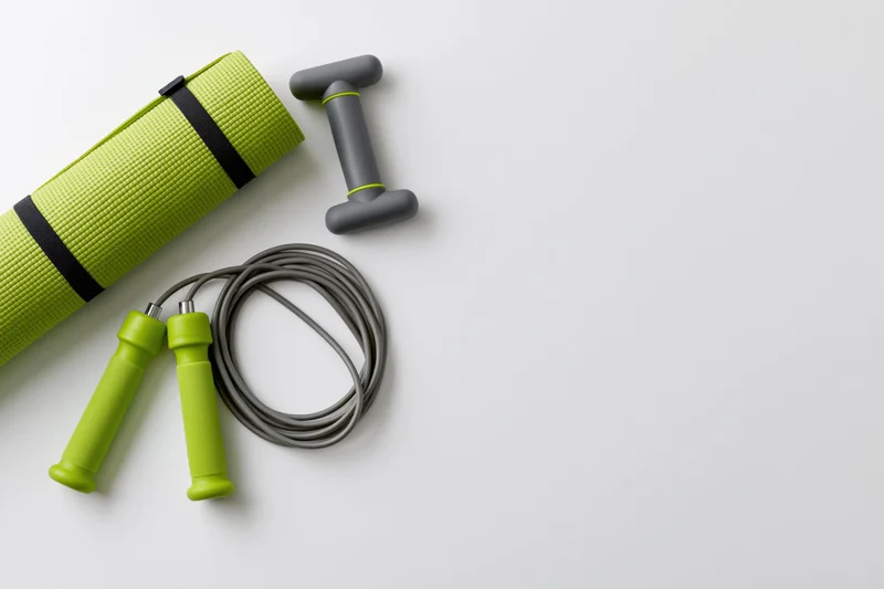 Rolled green yoga mat, gray dumbbell and jump rope arranged on white surface