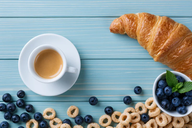 Flat lay breakfast spread with croissant, coffee, blueberries and cereal on light blue wooden table