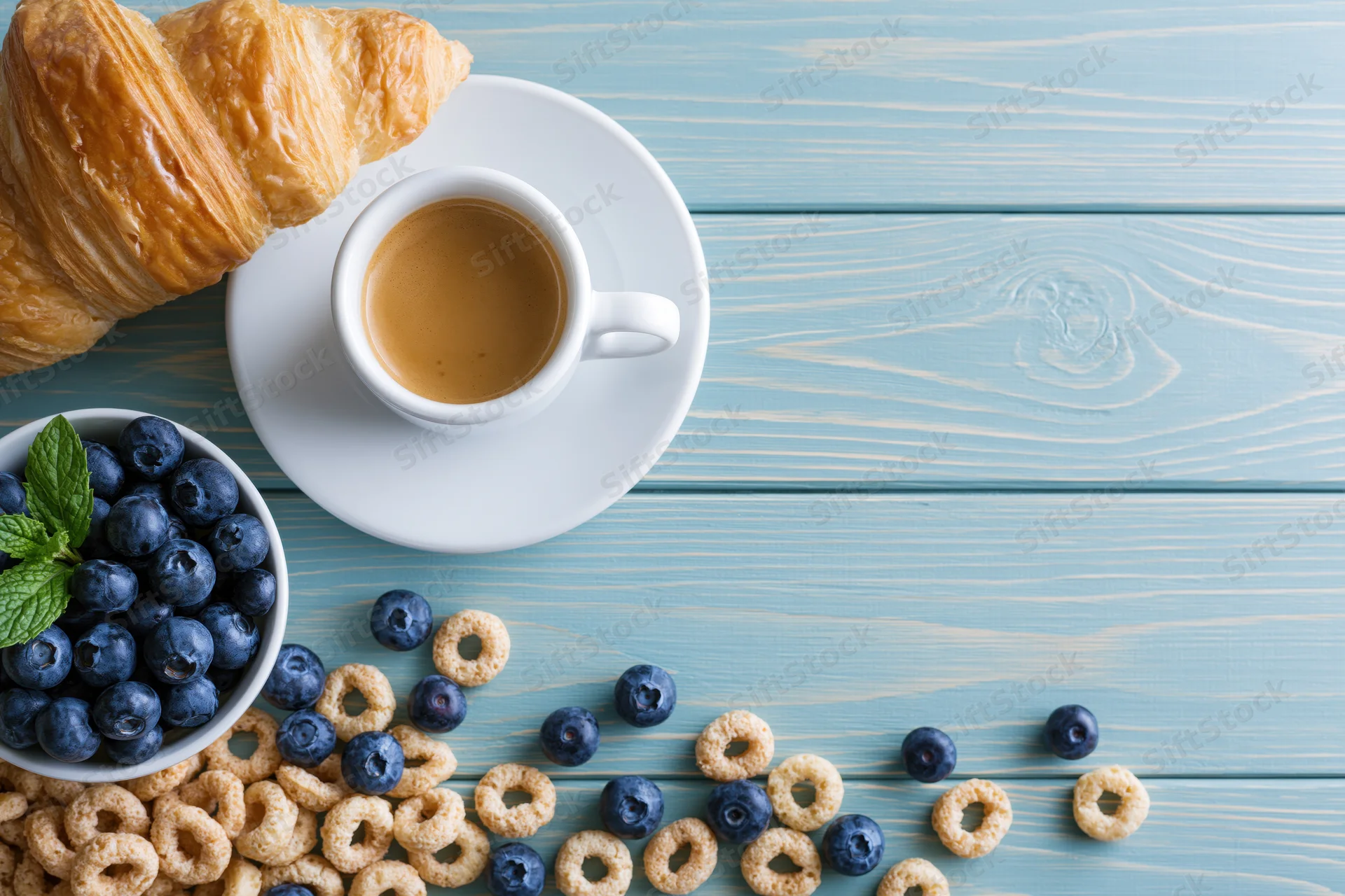 Breakfast spread with croissant, coffee, blueberries and cereal on light blue wooden table