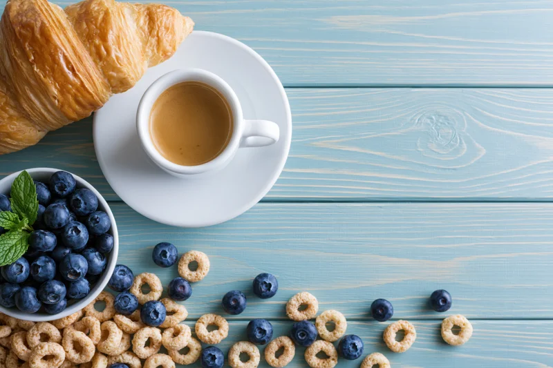 Breakfast spread with croissant, coffee, blueberries and cereal on light blue wooden table