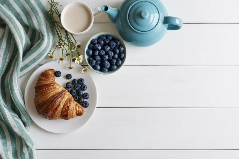 Flat lay breakfast with croissant, blueberries, and teapot on white wooden table