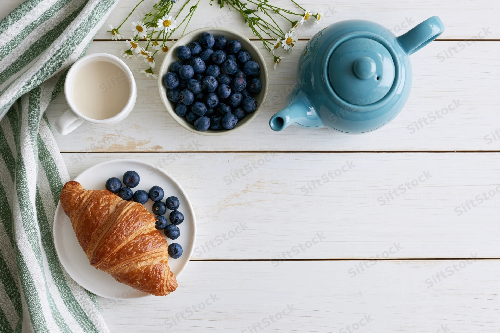Flat lay breakfast with croissant, blueberries, teapot and flowers on white wooden table