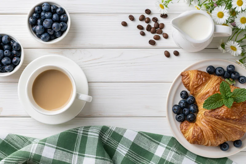 Breakfast spread with coffee, croissant, blueberries and milk on white wooden table