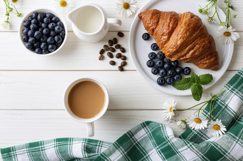 Flat lay breakfast spread with coffee, croissant, blueberries and daisies on white wooden table