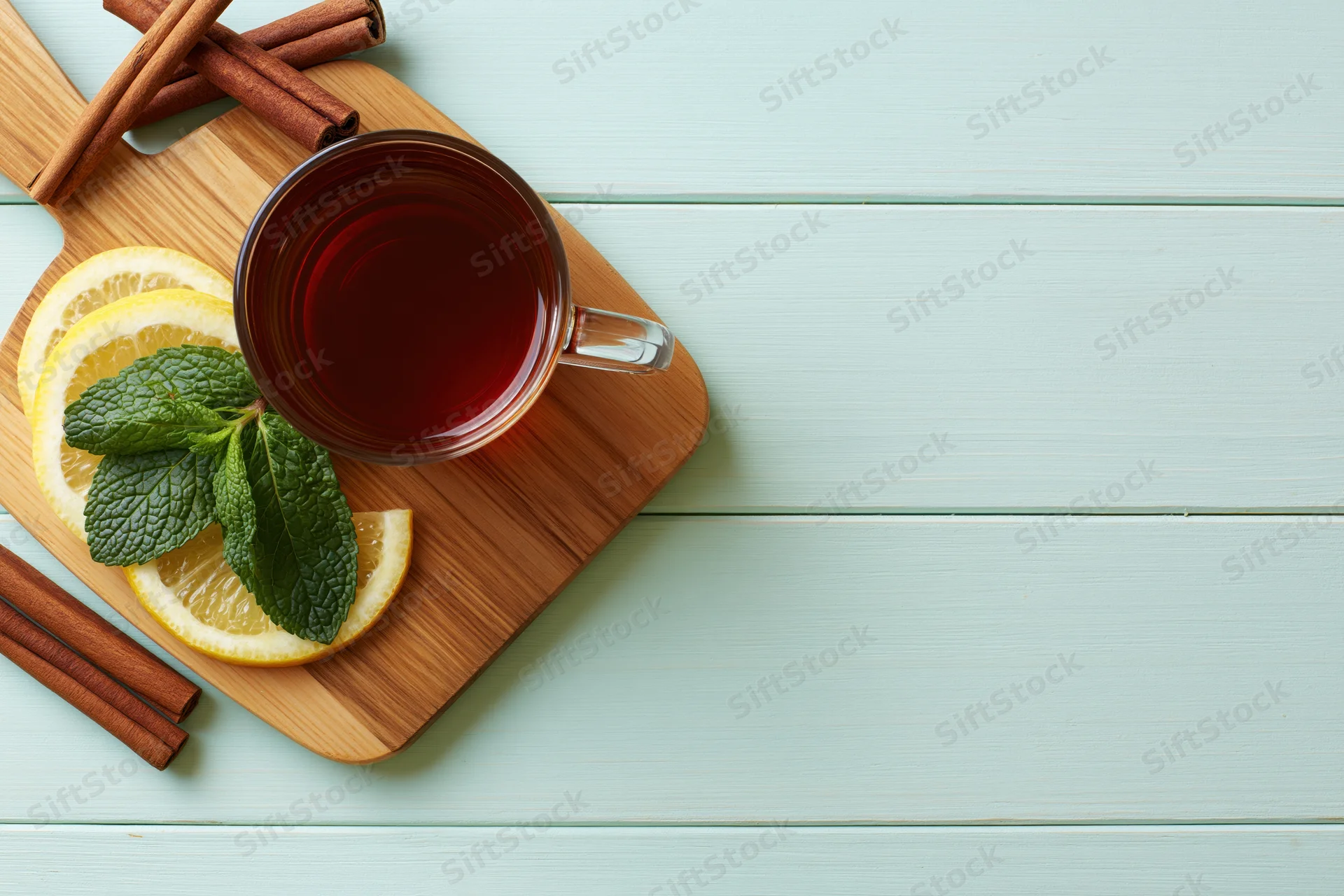 Glass mug of dark tea with lemon slices, mint leaves and cinnamon sticks on a wooden cutting board, placed on a light blue wooden surface, top view with copyspace
