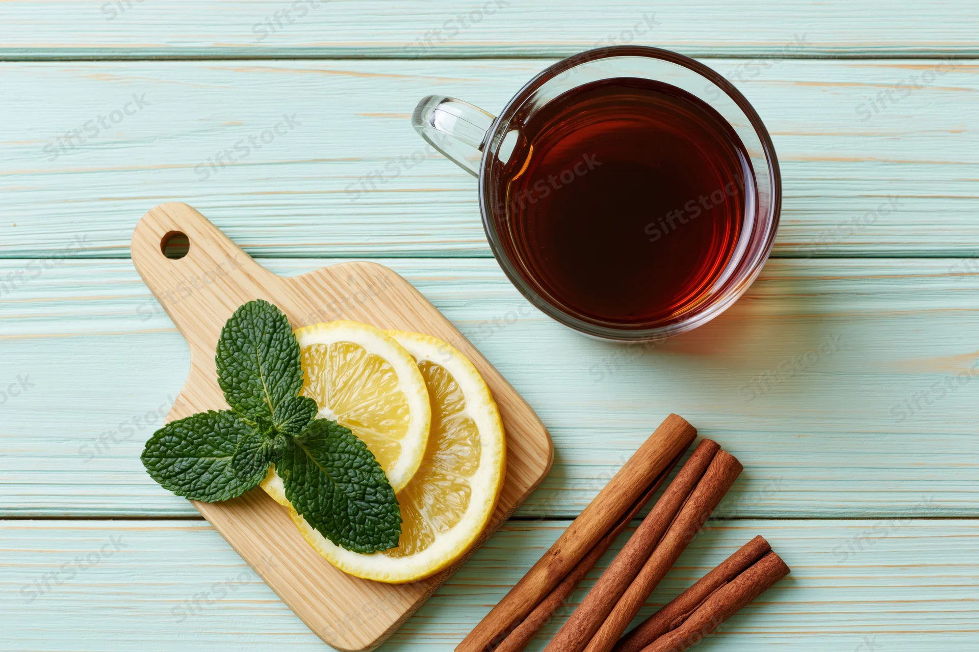 Glass mug of dark tea with lemon slices, mint leaves and cinnamon sticks on a wooden cutting board and light blue wooden surface