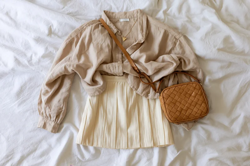Beige linen shirt layered over cream pleated skirt with brown woven handbag on white rumpled bedding