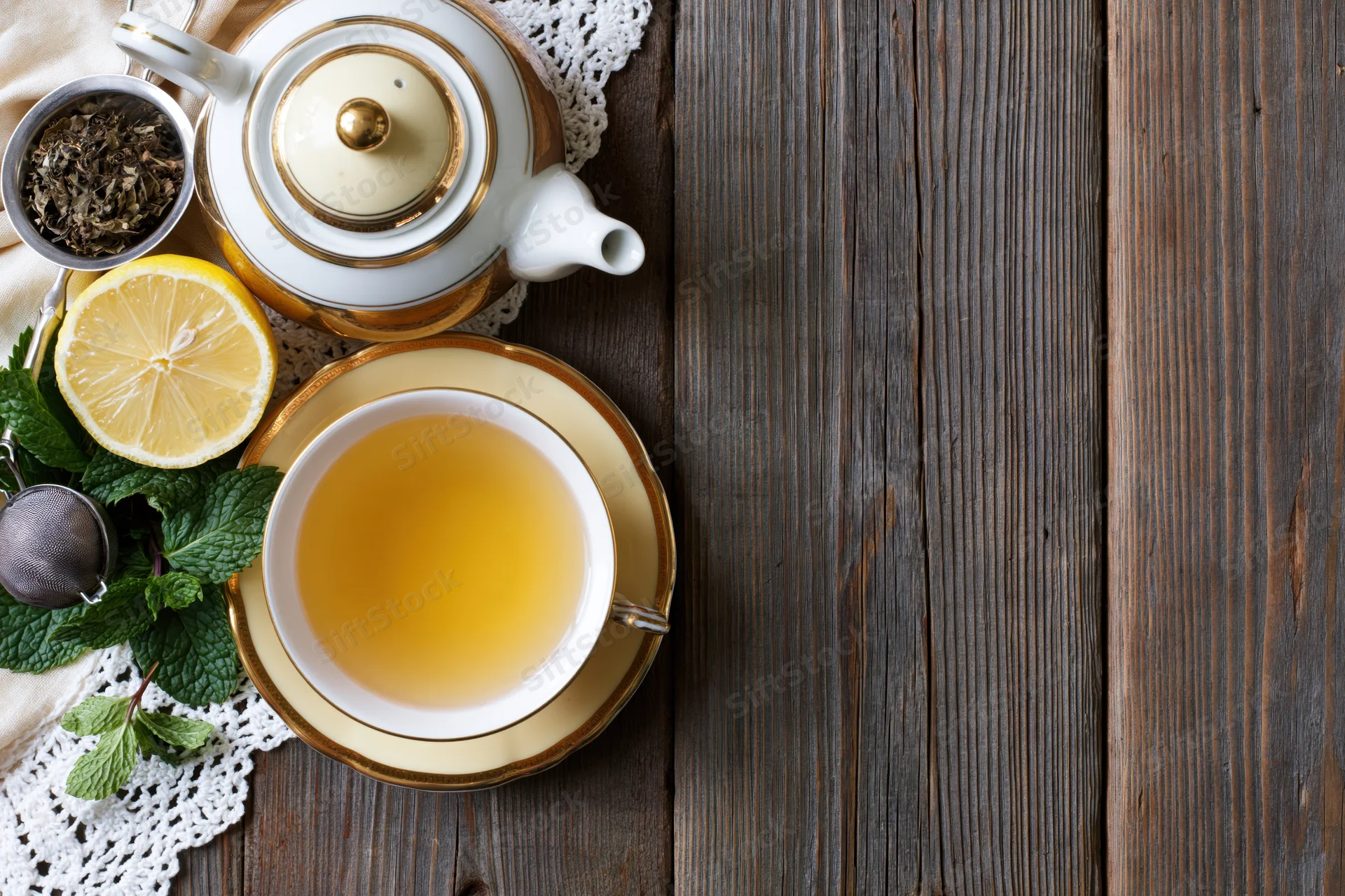 White and gold porcelain teapot and teacup with lemon, mint, and tea leaves on wooden table in flat lay arrangement