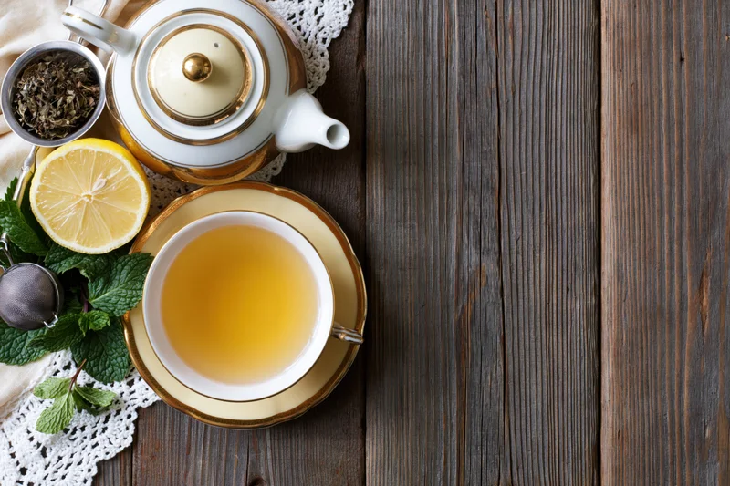 White and gold porcelain teapot and teacup with lemon, mint, and tea leaves on wooden table in flat lay arrangement