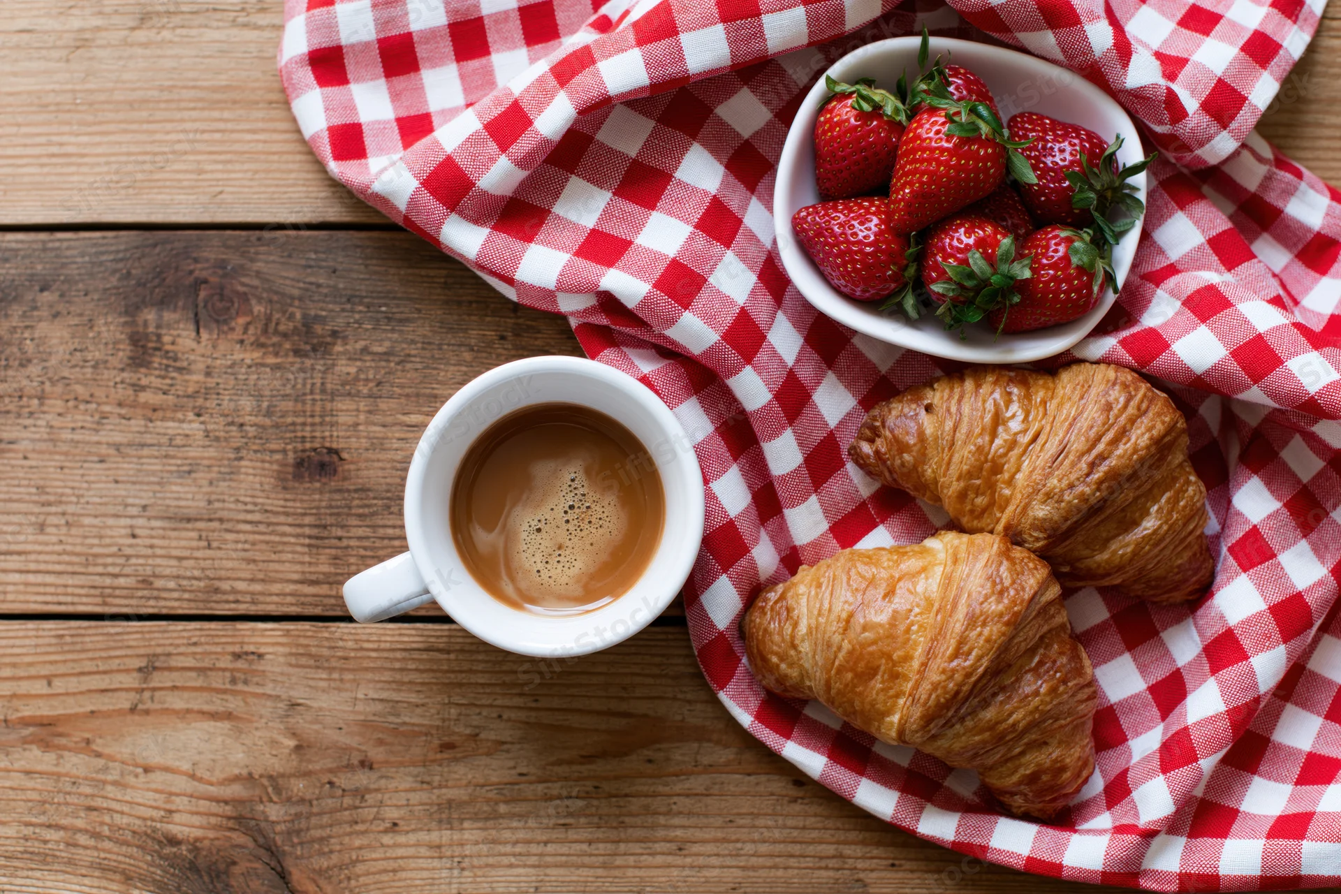 Breakfast flat lay with coffee, croissants and strawberries on a wooden table with red and white checkered cloth