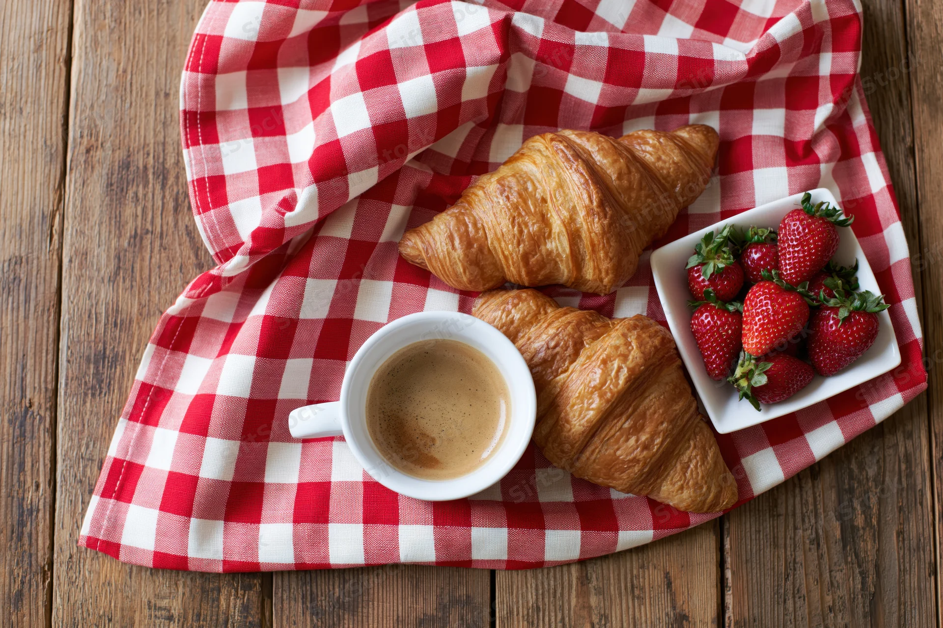 Two golden croissants, a cup of milky espresso, and a bowl of strawberries arranged on a red and white checkered cloth over a wooden table