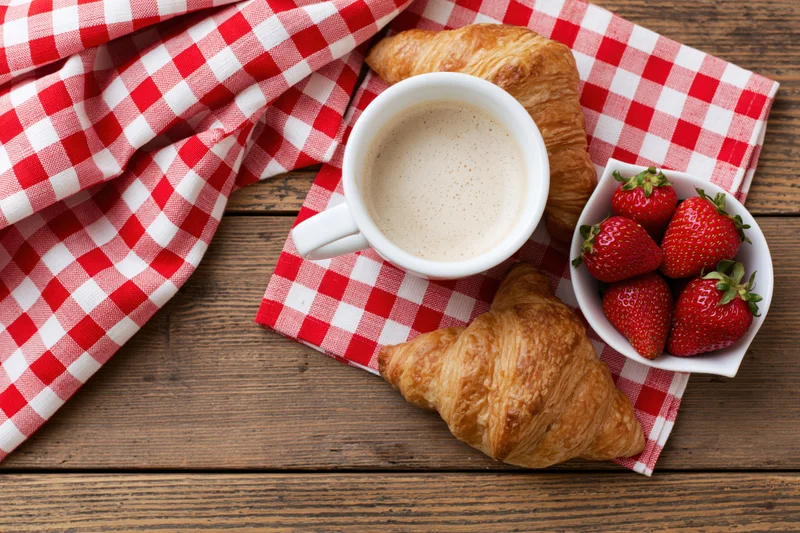 Flat lay of coffee cup with croissants and strawberries on checkered napkin over wooden table